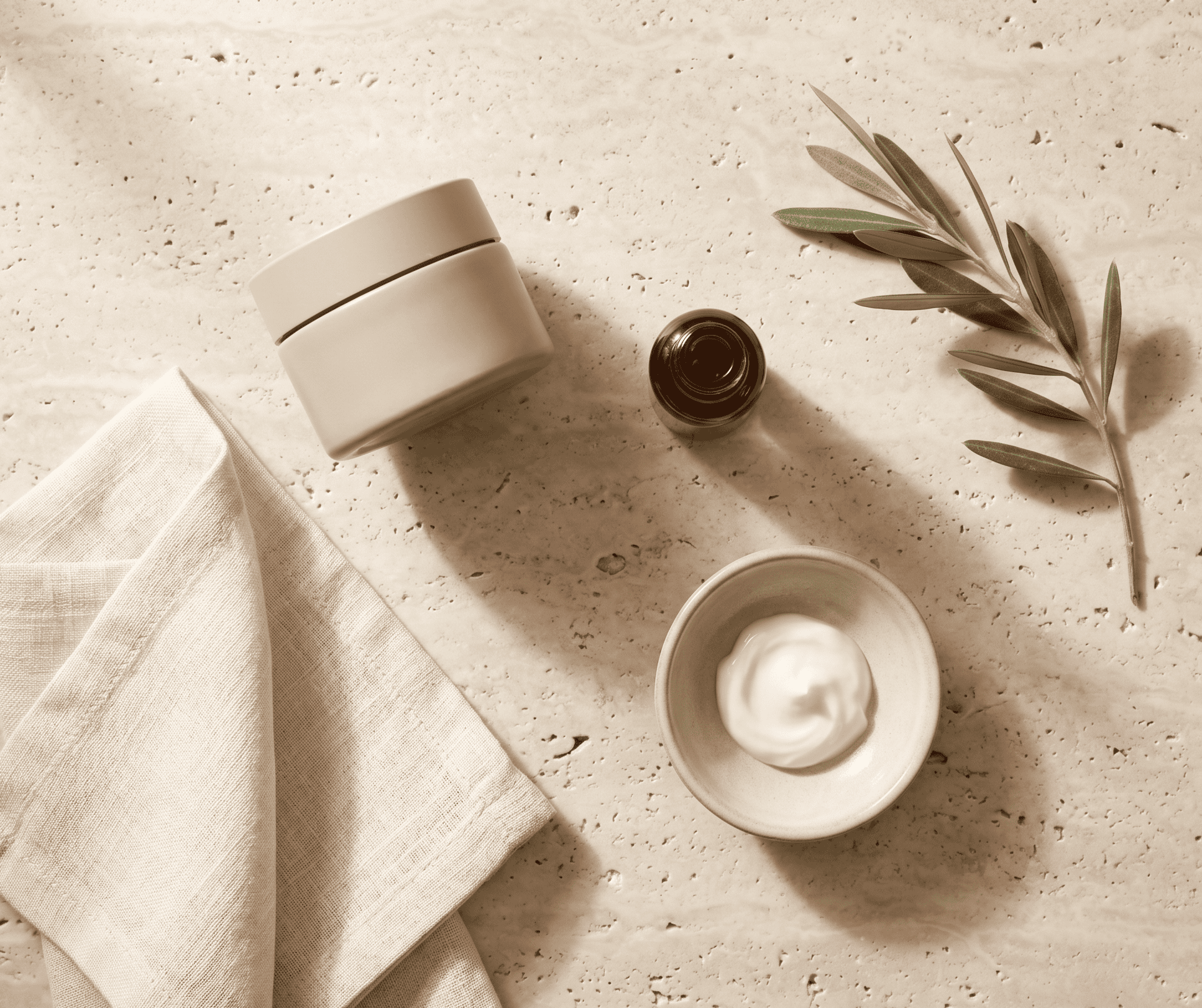 http://Crema%20hidratante,%20sérum%20y%20rama%20de%20olivo%20sobre%20mesa%20de%20mármol%20travertino%20en%20composición%20editorial%20cálida.