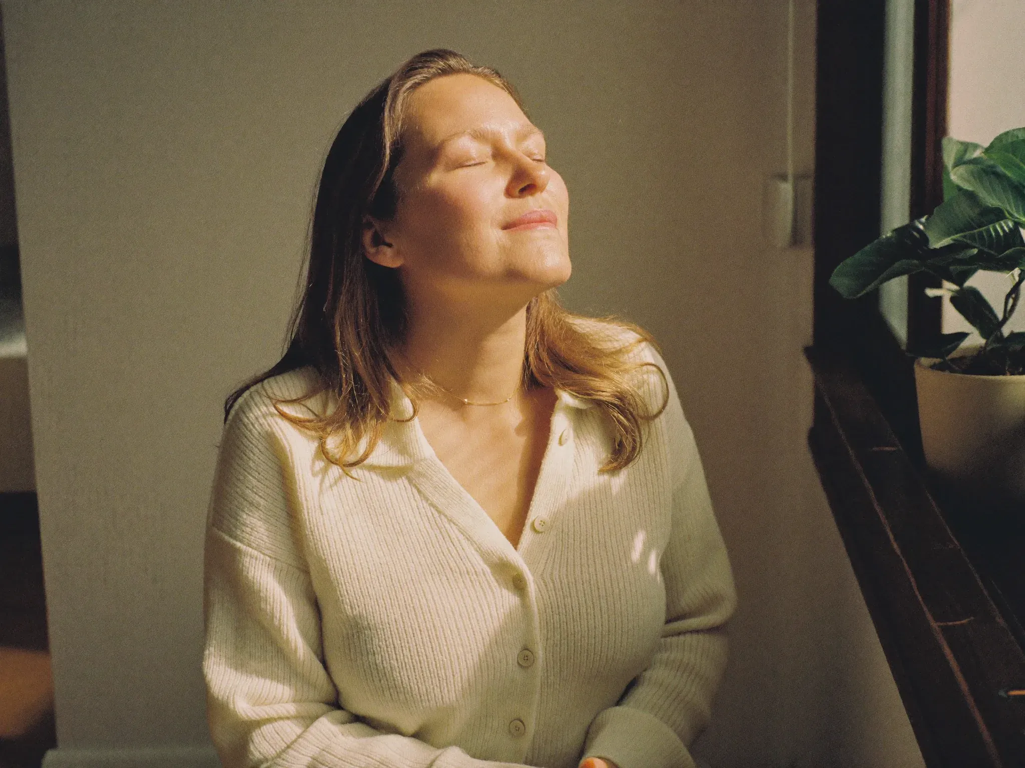 http://Mujer%20con%20los%20ojos%20cerrados%20recibiendo%20la%20luz%20del%20sol%20junto%20a%20la%20ventana,%20en%20calma.