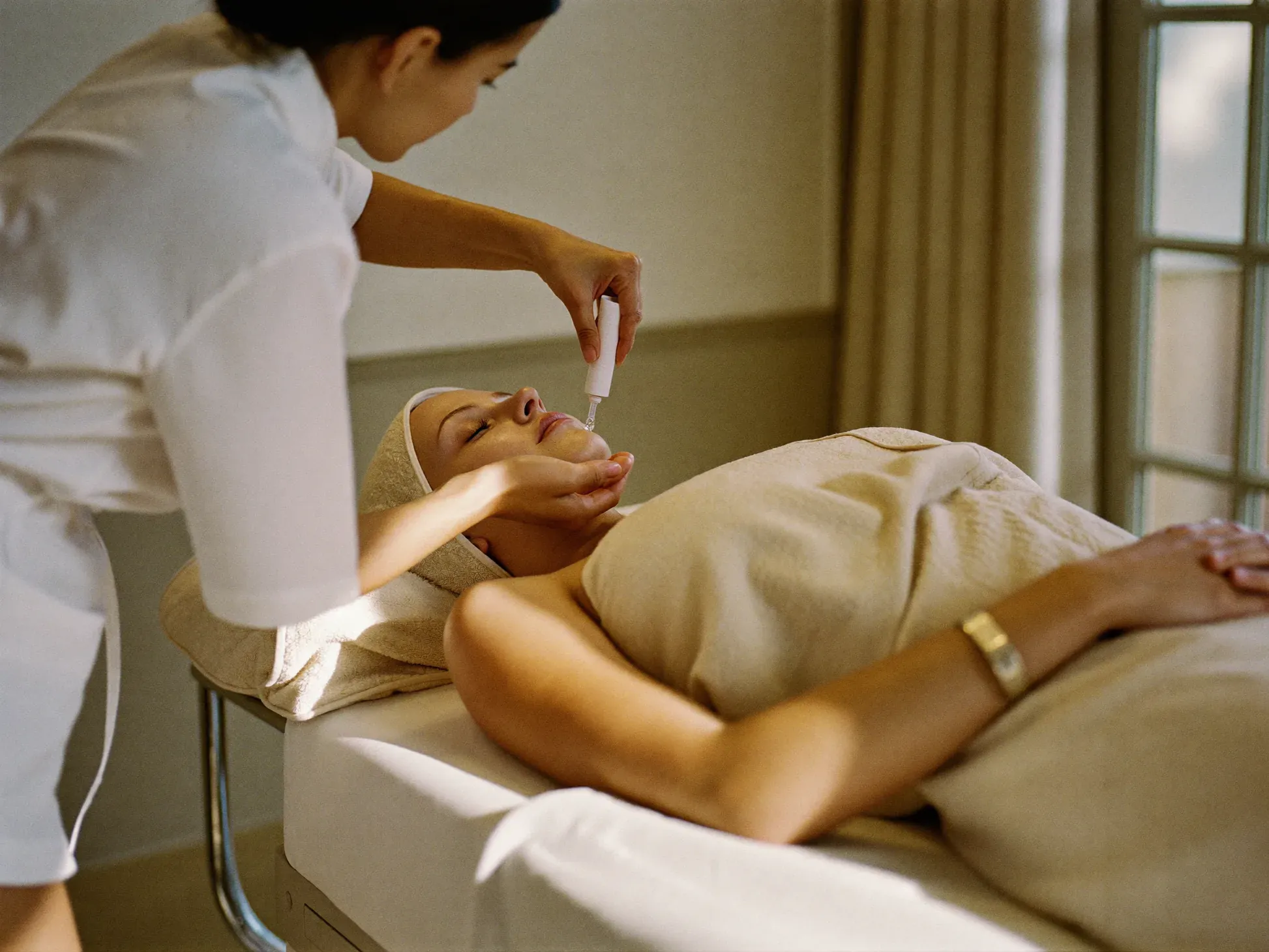 http://Especialista%20de%20NCA%20aplicando%20un%20sérum%20facial%20a%20una%20clienta%20recostada%20en%20la%20camilla%20de%20tratamiento.